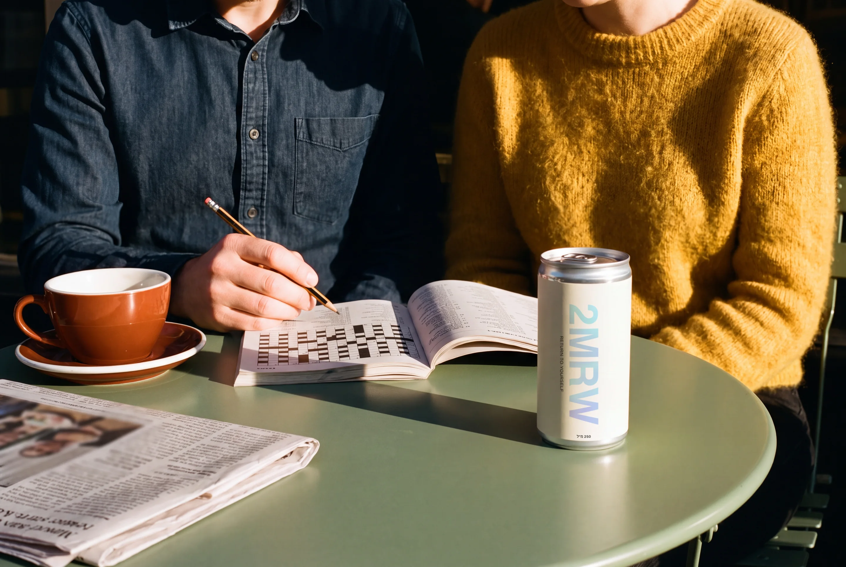 Two people at a green cafe table in low sun, one filling in a crossword, a 2MRW can standing in the foreground