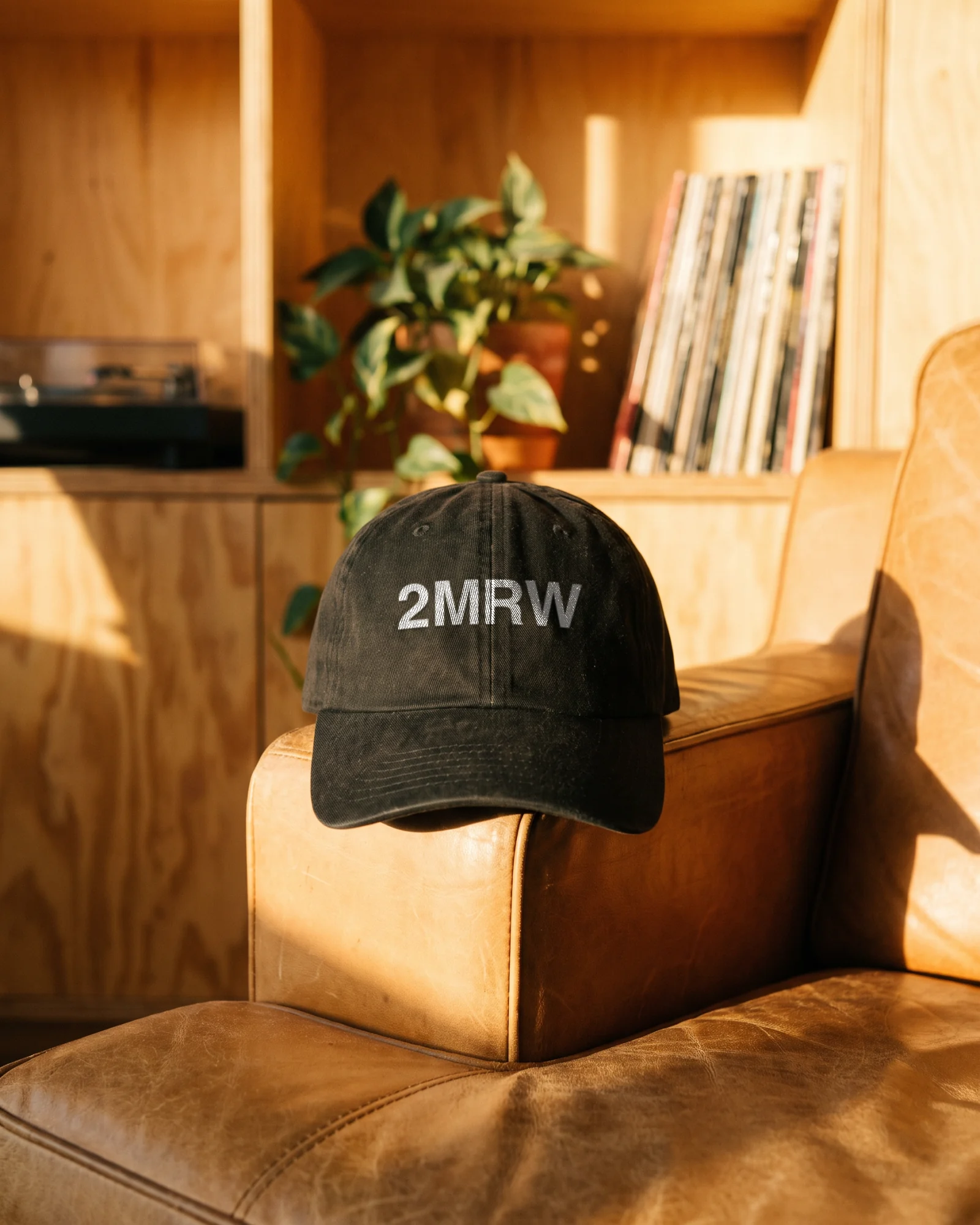 A washed black cap with the 2MRW wordmark resting on the arm of a tan leather chair