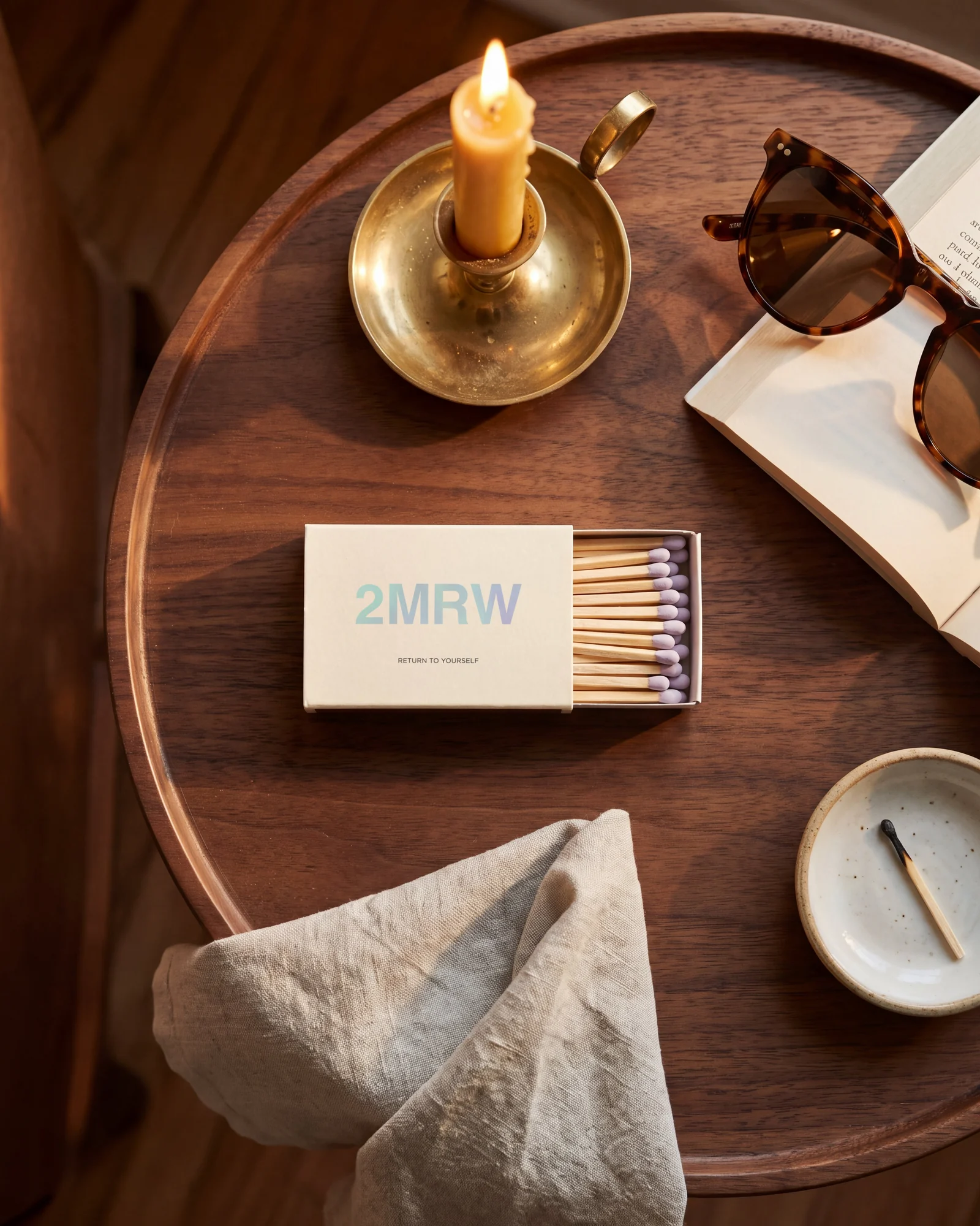 A small cream 2MRW matchbox on a walnut side table, its tray slid open to show lilac-headed matches, beside a lit candle and a pair of sunglasses