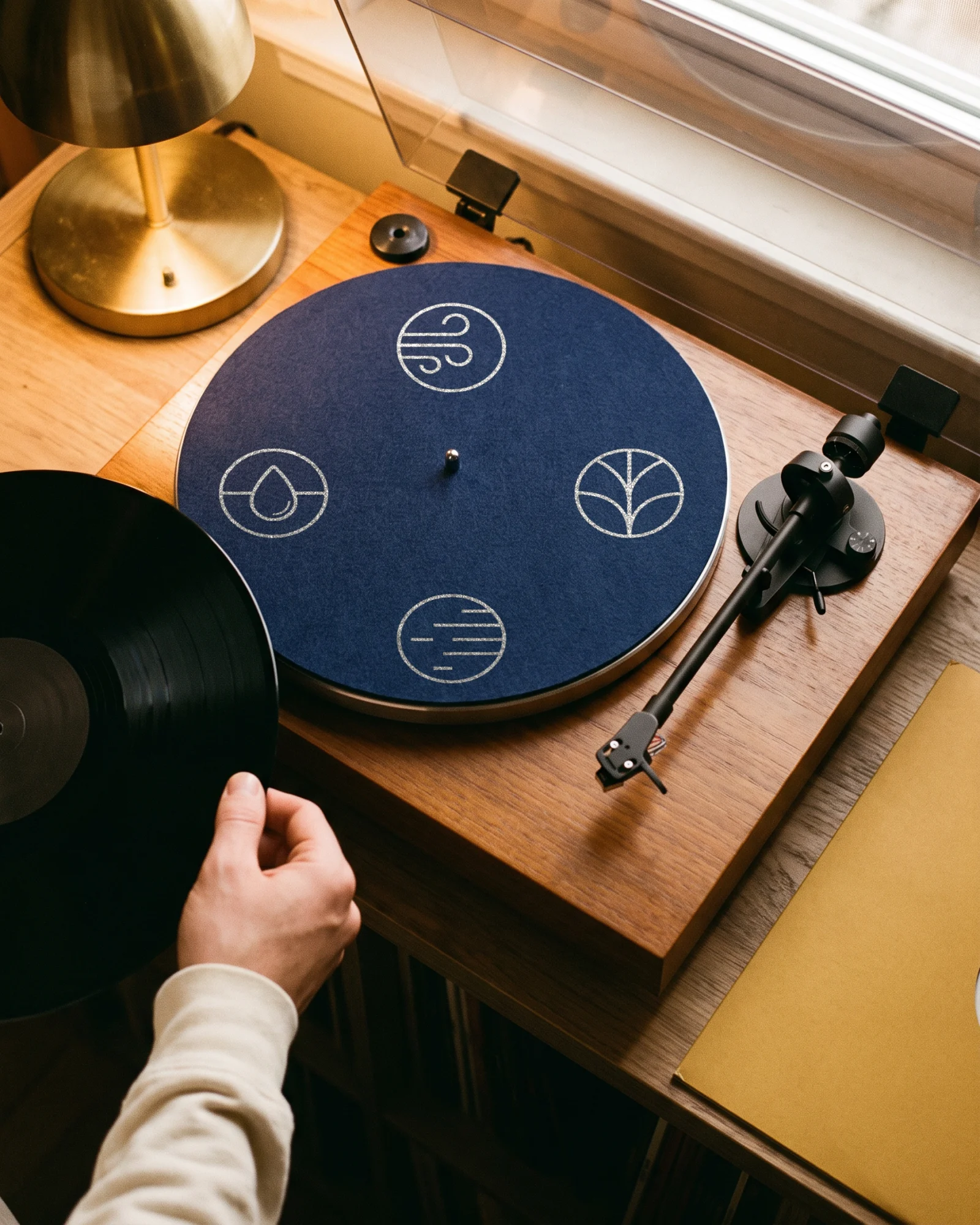 A turntable from above with a navy 2MRW slipmat printed with the four icons, a hand about to put a record on