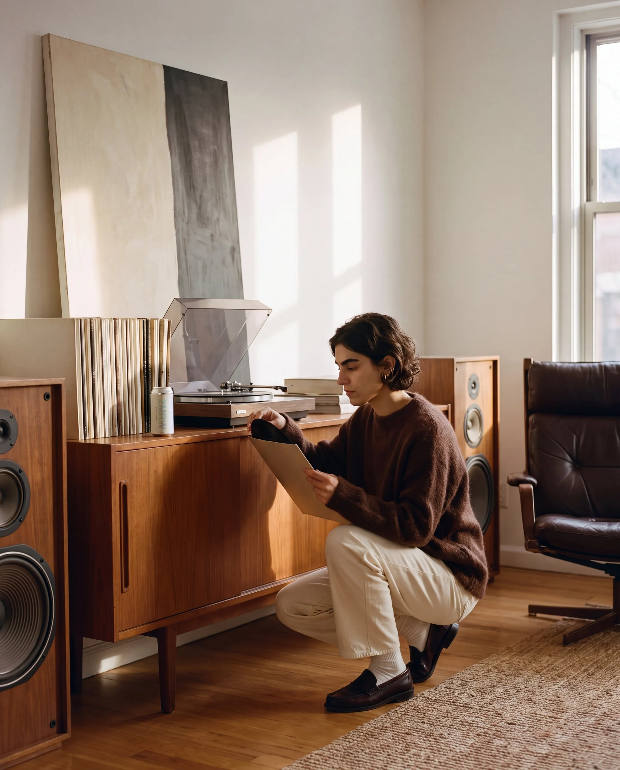 A woman in a brown mohair sweater crouches at a teak sideboard pulling a record from its sleeve, a 2MRW can beside the turntable