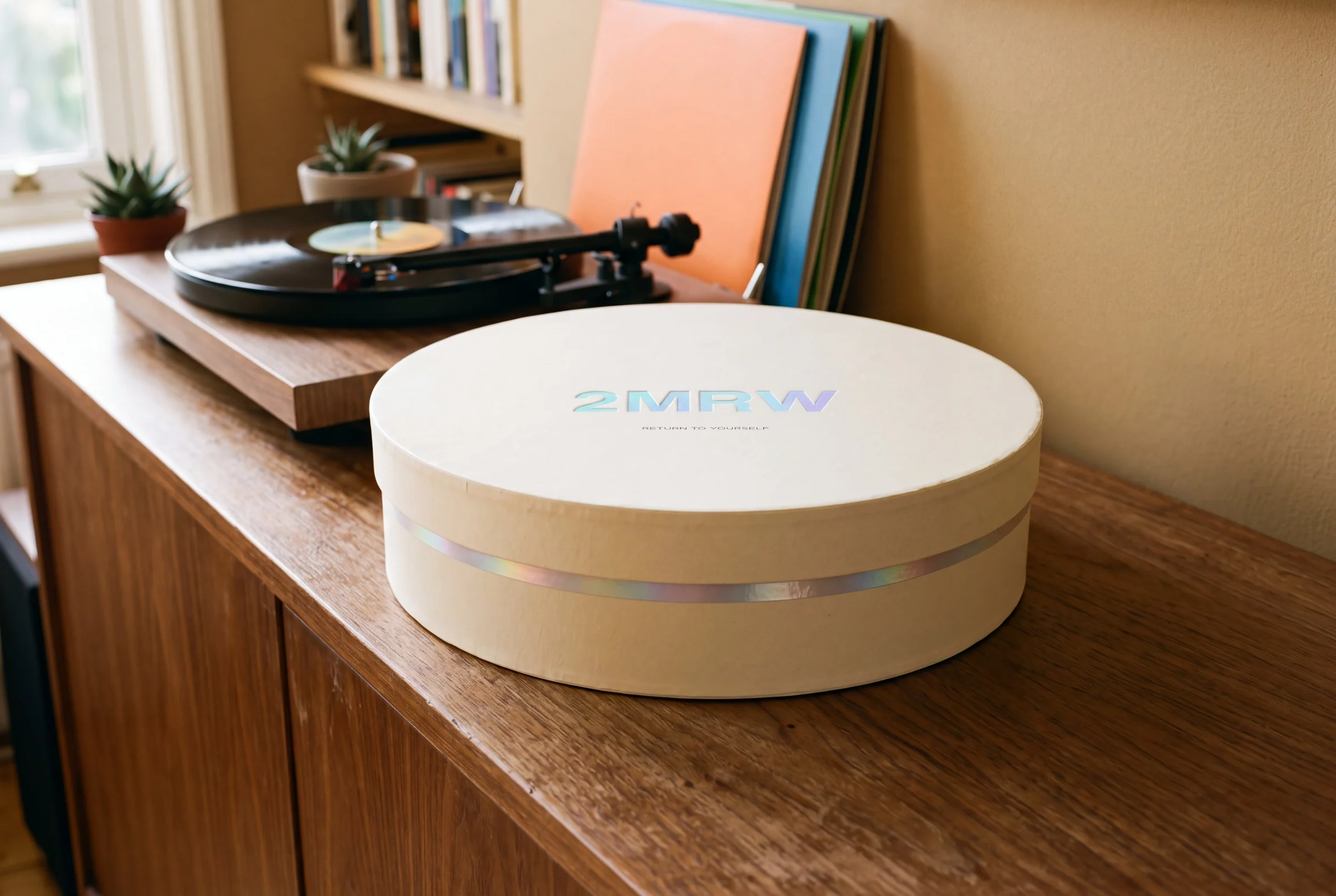 The closed round cream box on a sideboard beside a turntable: the 2MRW logo debossed in holographic foil on the lid and a thin line of foil where the lid meets the base