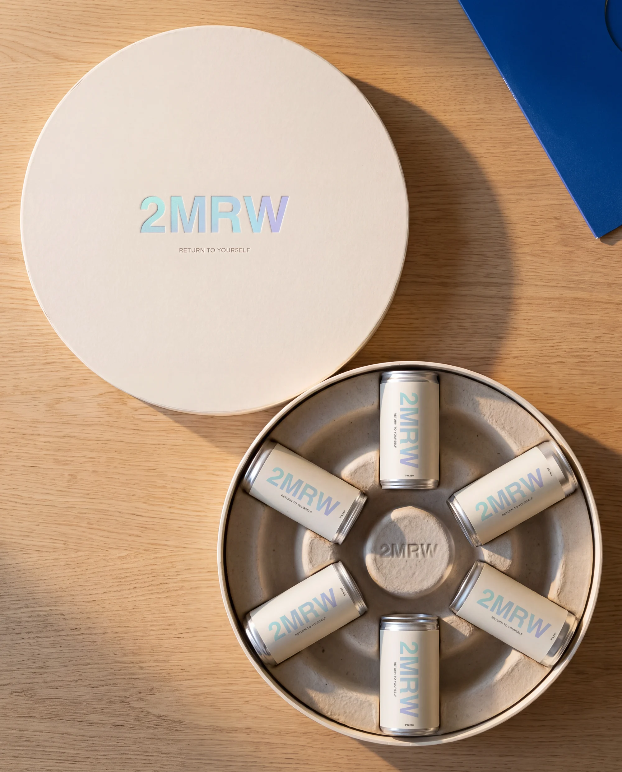 The round box opened on a pale oak table: six 2MRW cans lie in a grey moulded pulp tray like the spokes of a wheel, with 2MRW pressed into the pulp at the centre. The cream lid lies beside it with the logo debossed in holographic foil