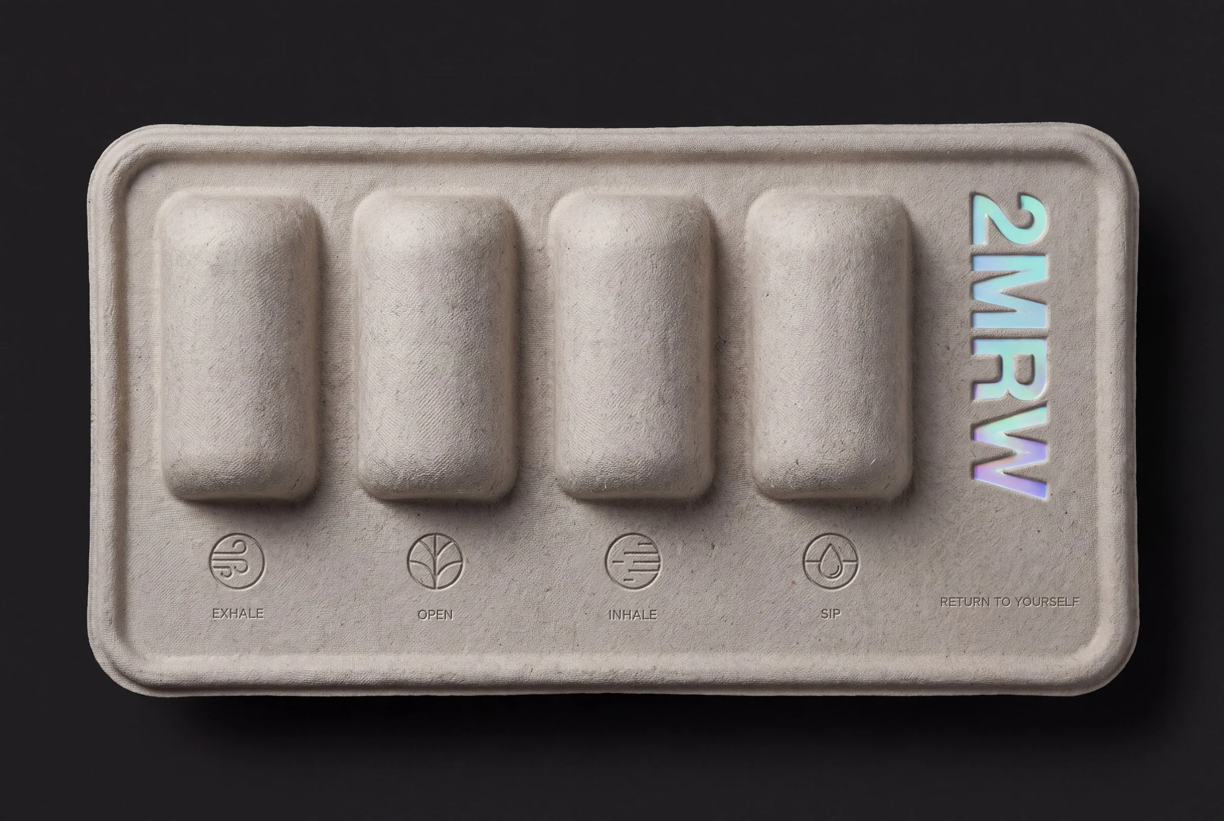 A grey moulded pulp pack on black: four raised can forms in a row, the four ritual icons and words pressed into the pulp beneath them, and the 2MRW logo debossed in holographic foil up the right side
