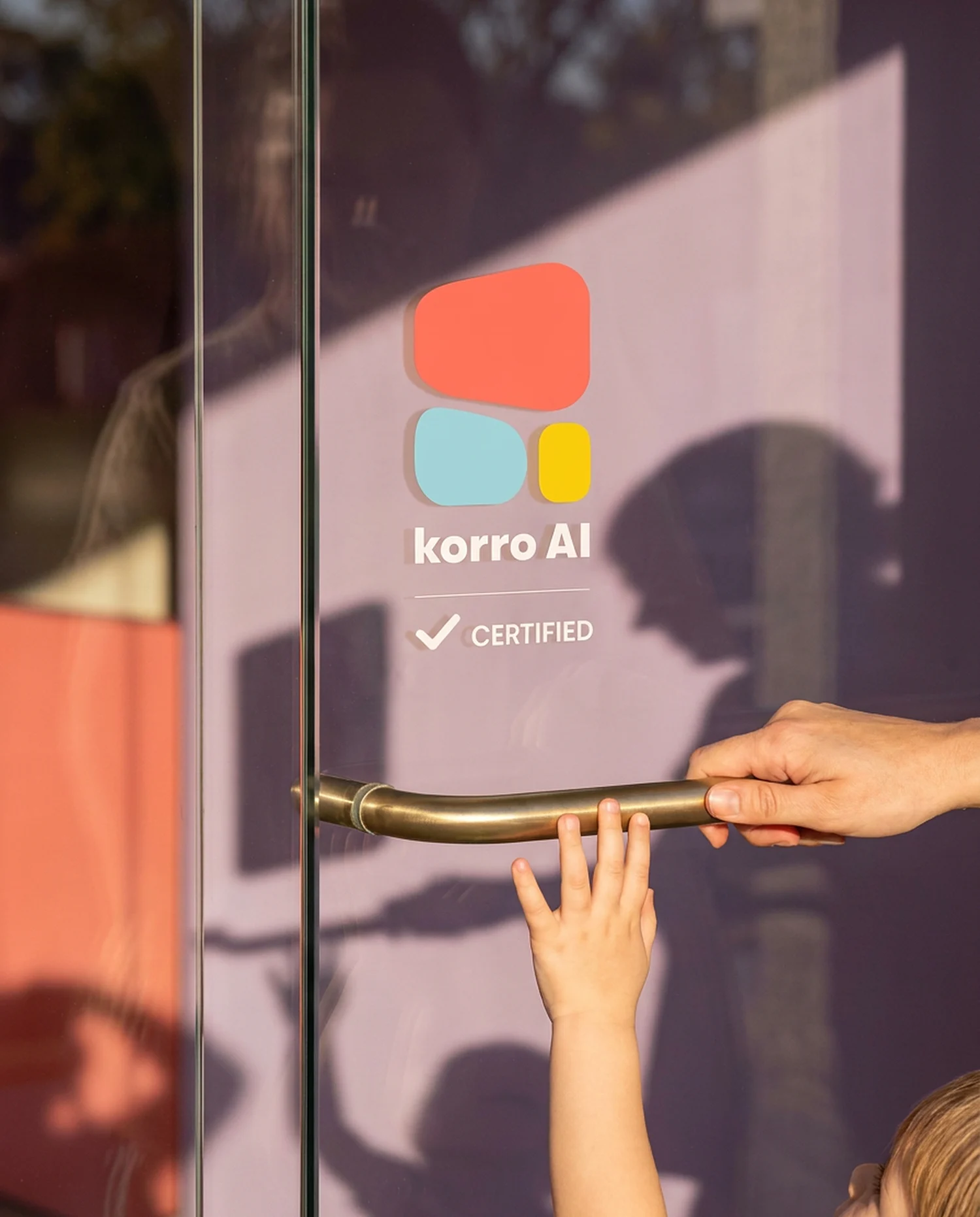 The Korro AI Certified badge as a clear decal on a glass clinic door, the coral, blue and yellow symbol and white lettering straight on the glass with a lilac wall behind, an adult hand on the brass handle and a child's hand reaching up