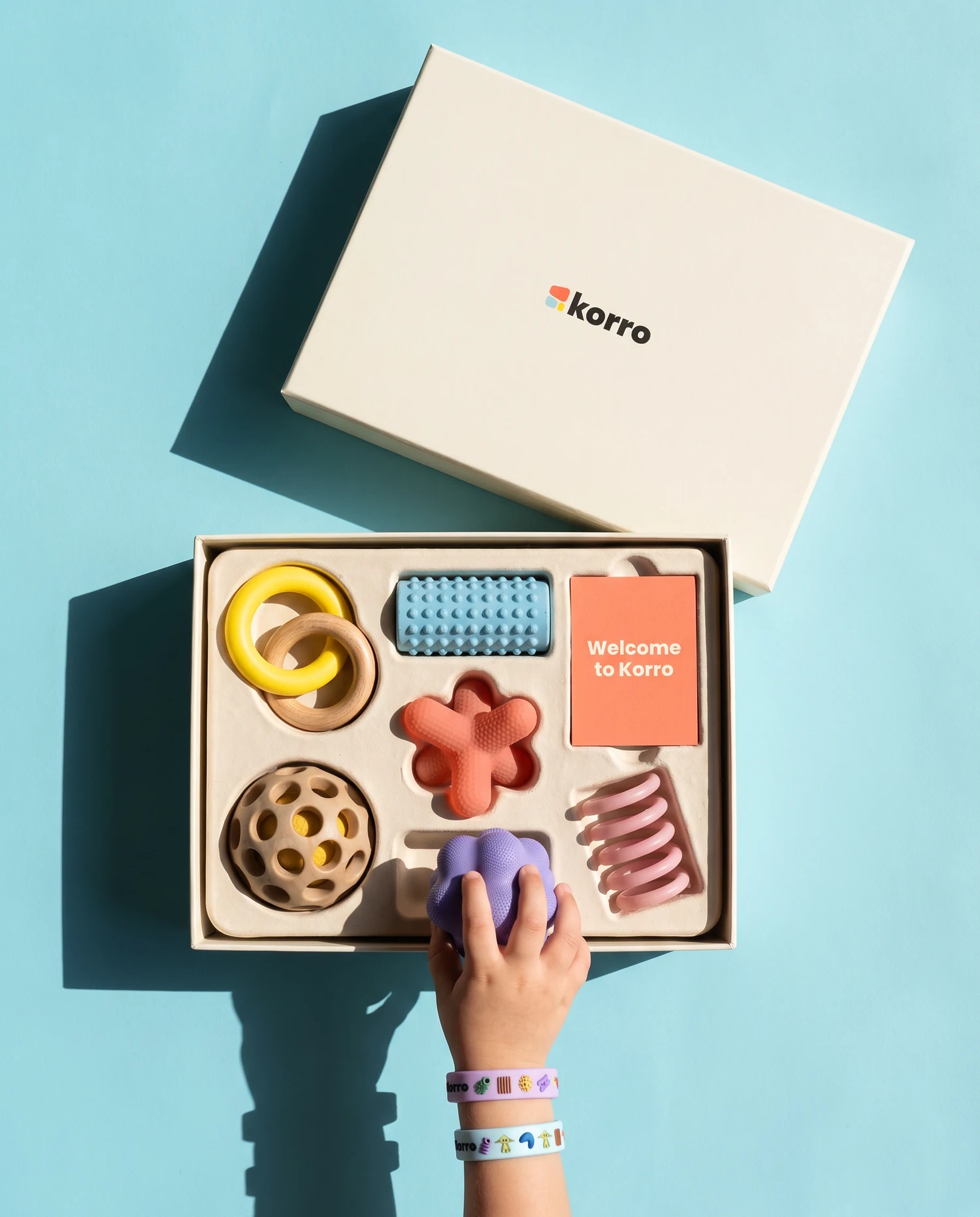 Seen from above on sky-blue paper in hard sunlight: an open cream Korro box with a moulded tray holding linked yellow and wooden rings, a blue studded roller, a coral jack, a wooden honeycomb ball, a pink coil and a card reading welcome to Korro; the lid with the Korro logo lies beside it and a child's hand wearing two Korro wristbands lifts a lilac form out