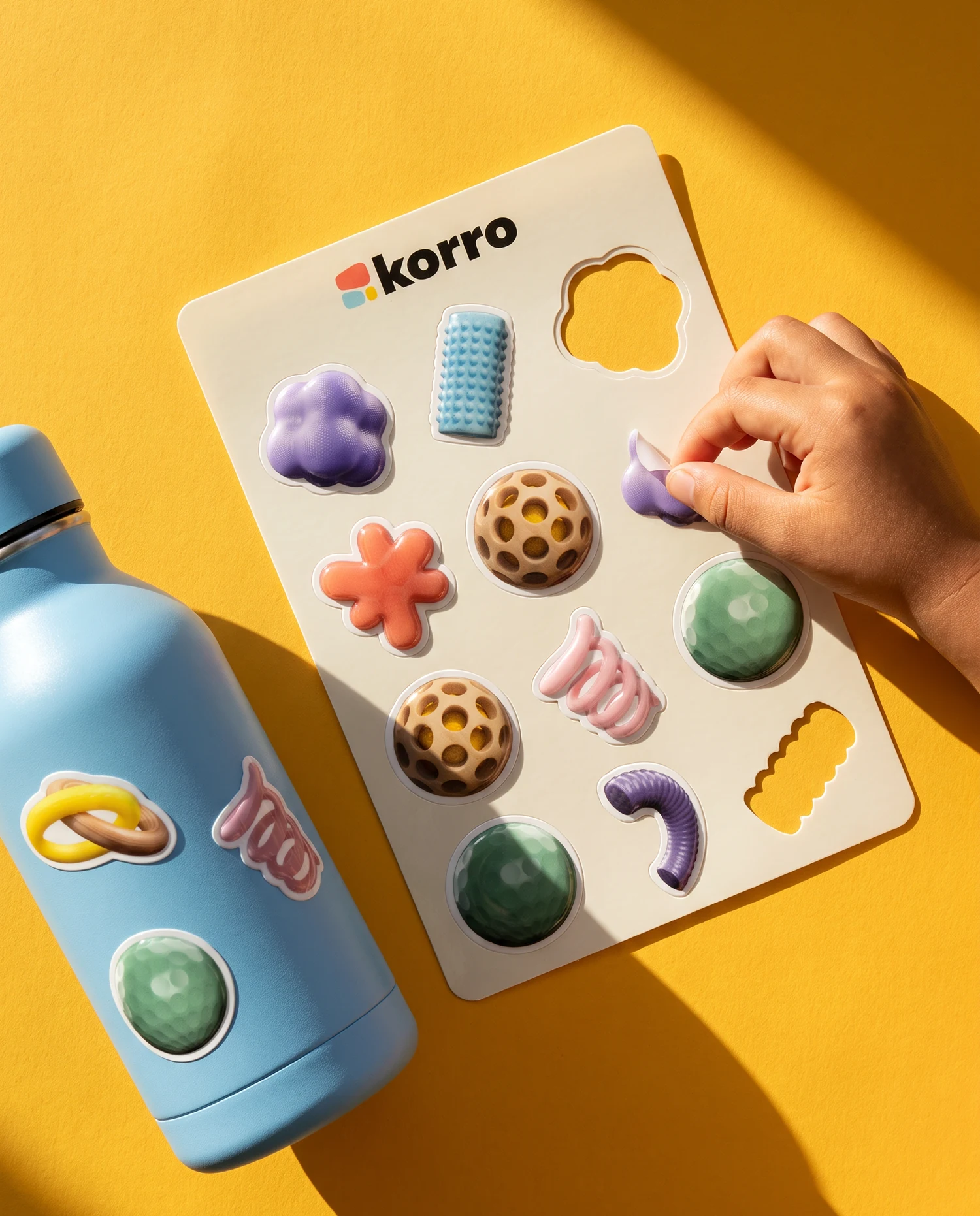 On yellow paper in hard sunlight, a cream sticker sheet with the Korro logo and puffy stickers of the forms, a child's hand peeling one off, and a sky-blue water bottle with three of the stickers on it