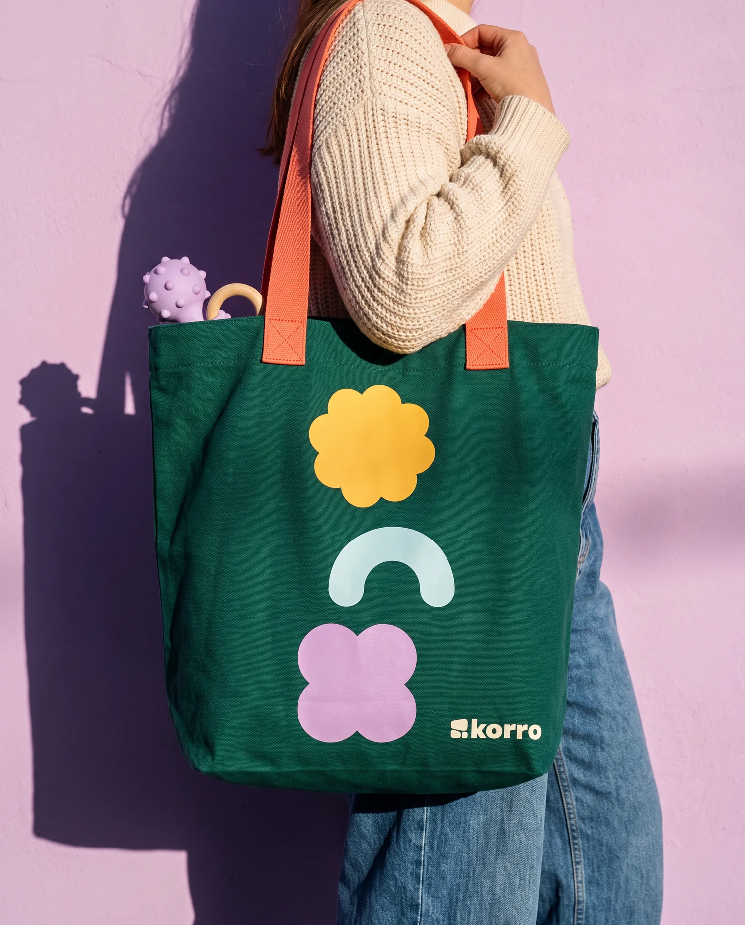 A deep green canvas tote with coral straps on a shoulder, printed with a yellow flower, a pale blue arch and a lilac clover, against a sunlit lilac wall