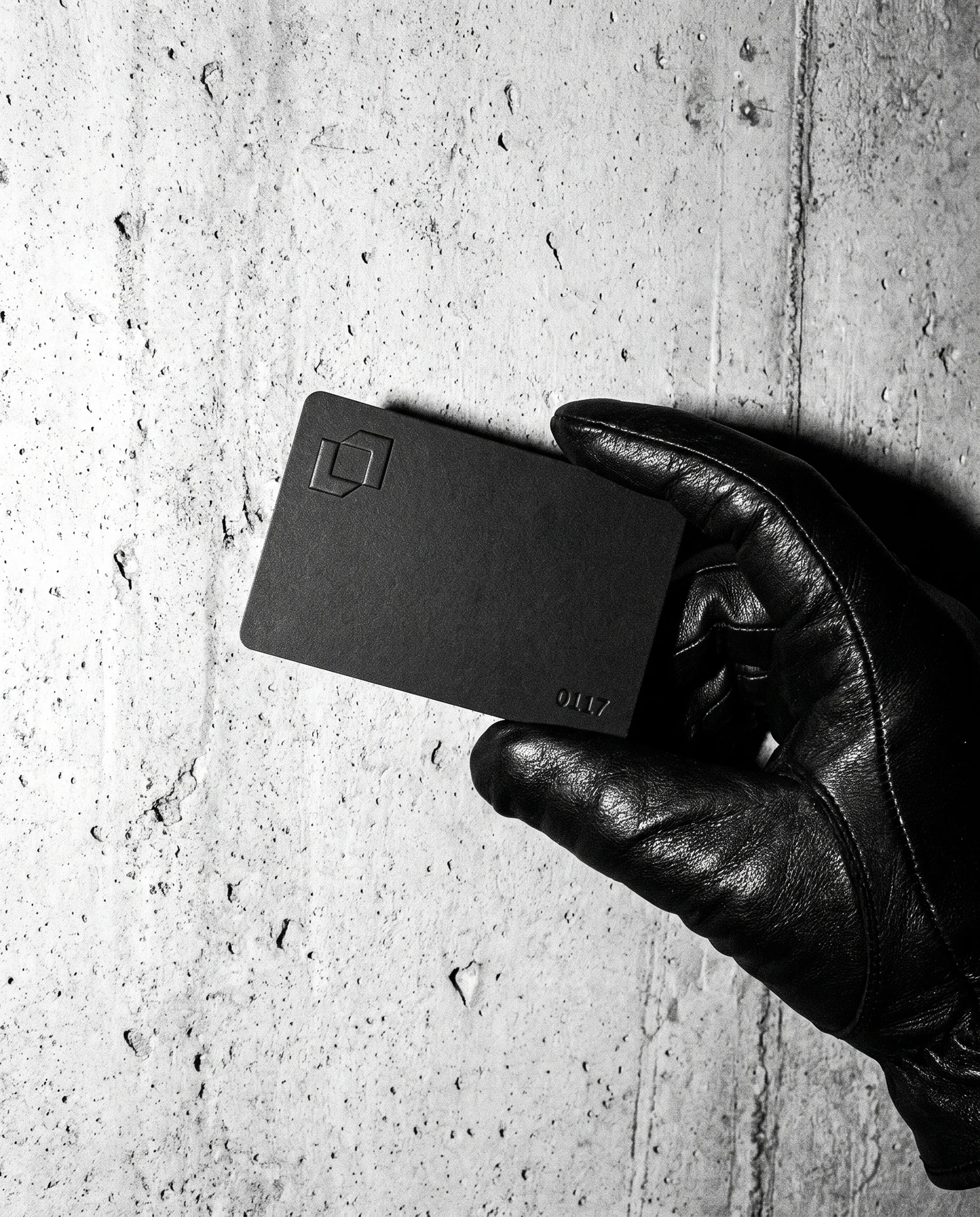 A gloved hand holding a black card against a concrete wall, the symbol embossed small in the corner