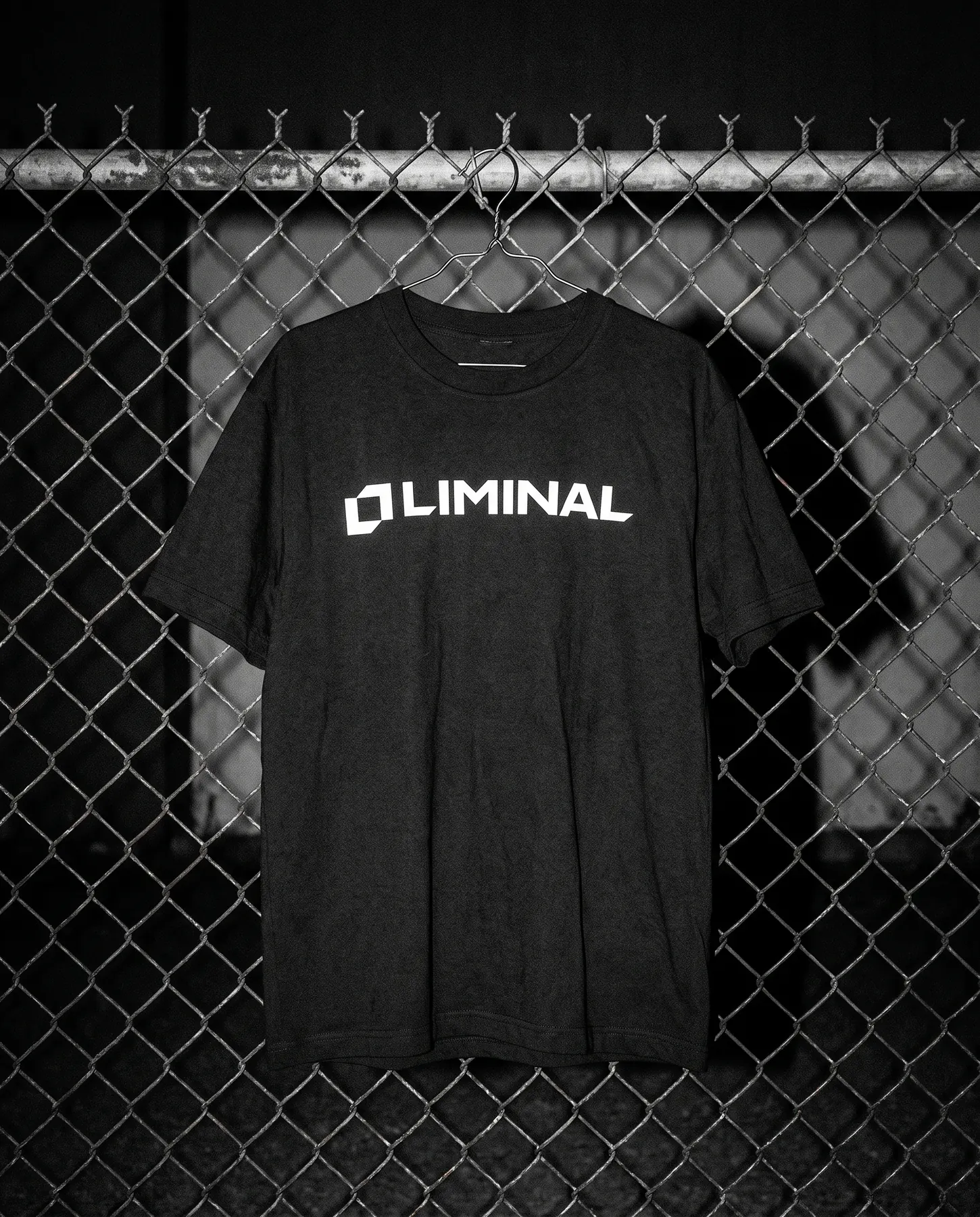 Black t-shirt with the Liminal lockup on a wire hanger on a chain-link fence, shot with flash