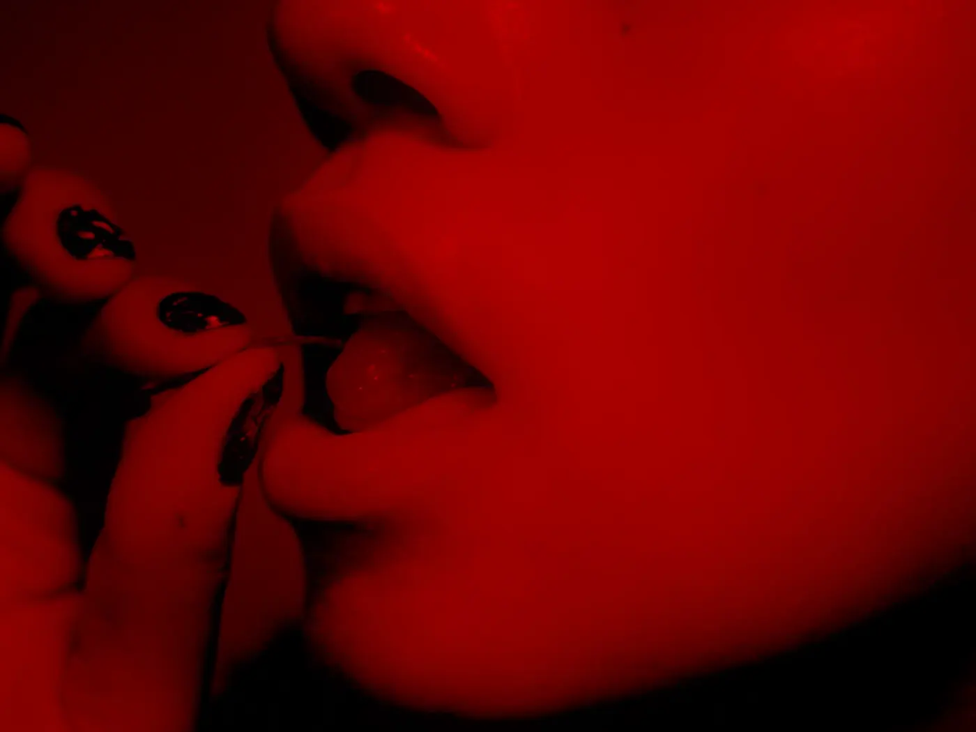 A cocktail cherry held to a mouth by a hand with painted nails, in deep red light