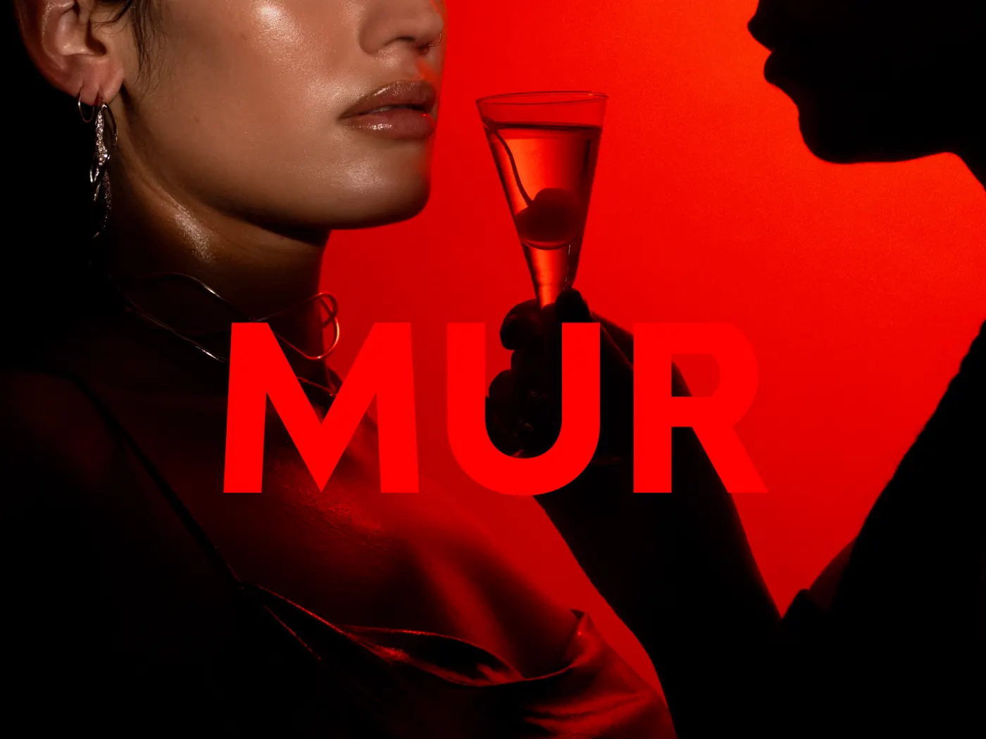 A woman holding a coupe against a red wall, a silhouetted profile facing her from the other side, with MUR set across the middle in red