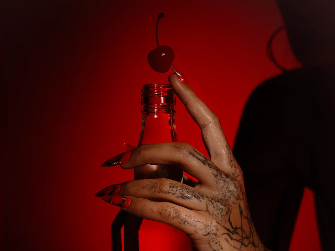 A tattooed hand holding a Sok&oacute;ł bottle with a cocktail cherry balanced on the rim, lit hard red