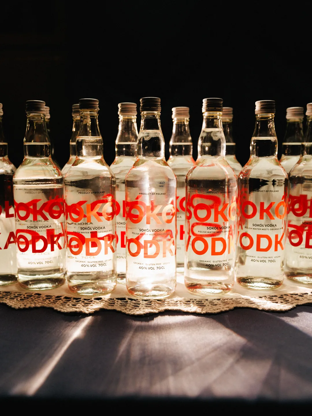 A row of Sok&oacute;ł bottles on a lace runner, the oversized red wordmark on each one cropped differently by the curve of the glass