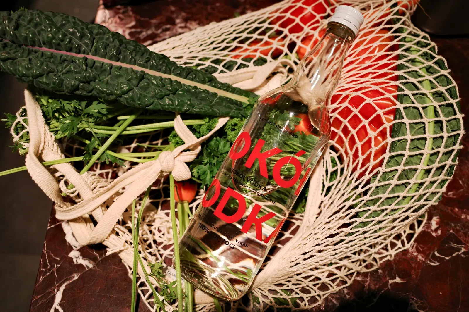 A Sok&oacute;ł bottle lying in a string shopping bag with kale, parsley and tomatoes on a marble counter