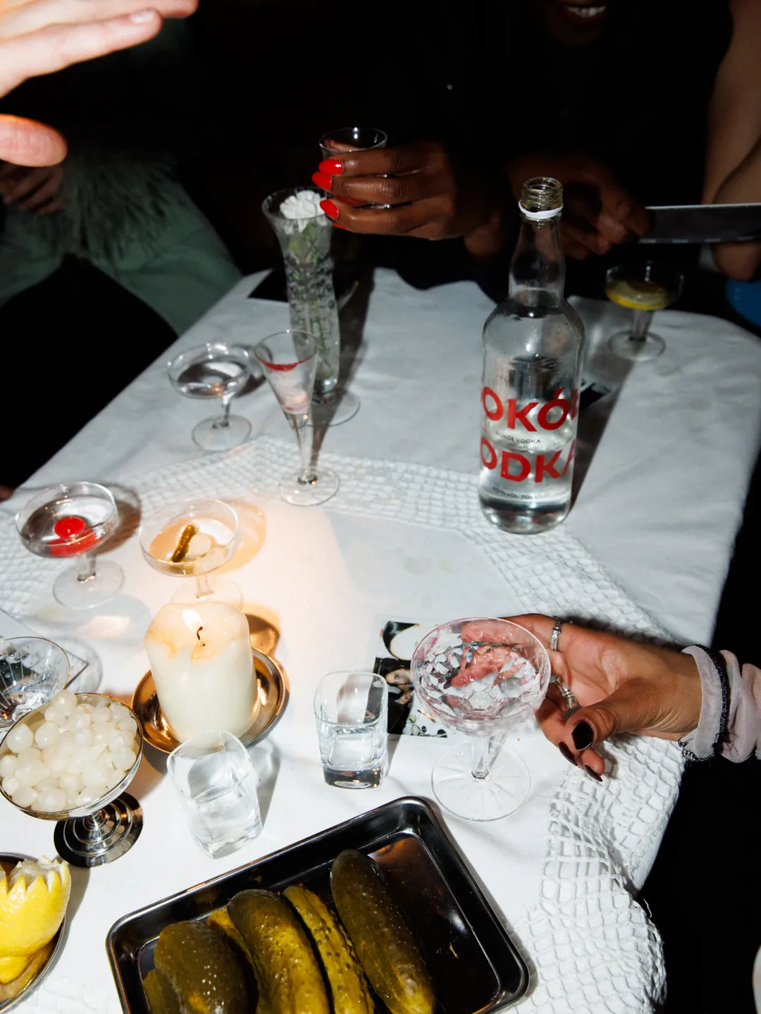 A supper table photographed from above: pickles, cocktail onions, lemons, a burning candle, coupes and hands reaching in