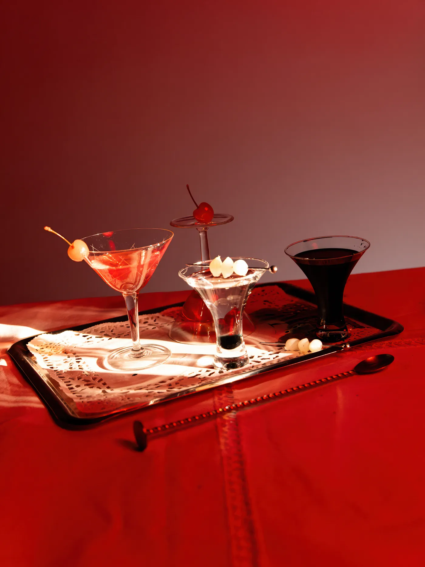 Three cocktails on a silver tray on a red tablecloth, lit by a hard shaft of light
